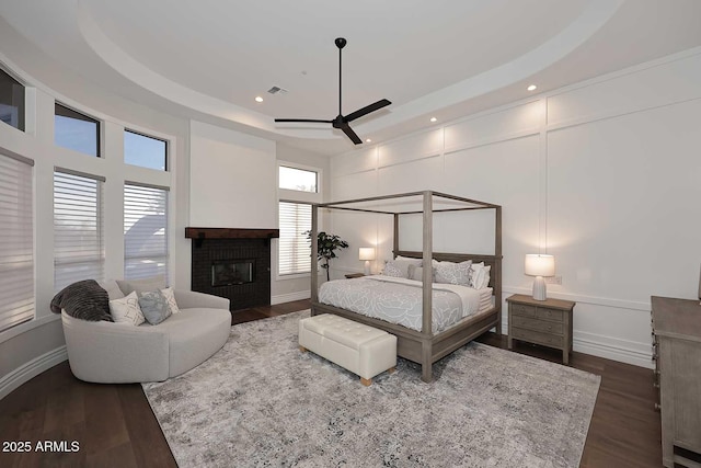 bedroom featuring a brick fireplace, dark hardwood / wood-style flooring, ceiling fan, and a raised ceiling