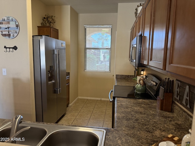 kitchen featuring light tile patterned flooring, dark countertops, appliances with stainless steel finishes, a sink, and brown cabinetry
