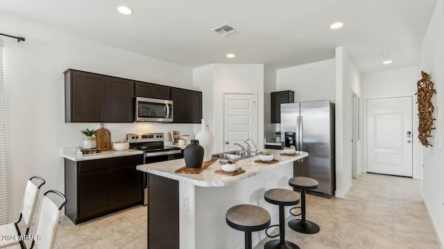 kitchen with sink, dark brown cabinets, appliances with stainless steel finishes, a kitchen breakfast bar, and an island with sink