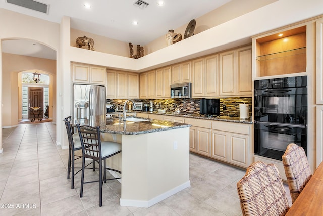 kitchen featuring sink, a kitchen island with sink, dark stone counters, appliances with stainless steel finishes, and light brown cabinets