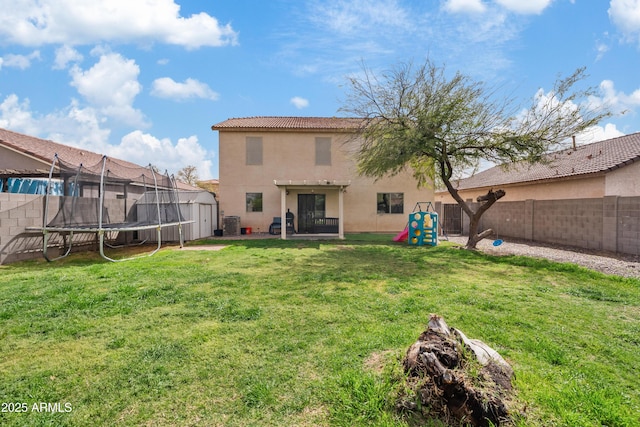 back of house with stucco siding, a lawn, a tile roof, a trampoline, and a fenced backyard