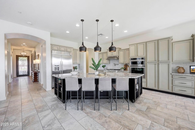 kitchen with appliances with stainless steel finishes, light stone countertops, a kitchen island with sink, and hanging light fixtures