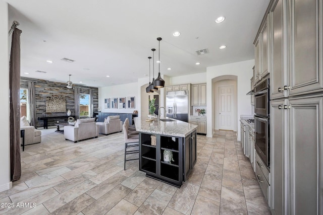 kitchen with sink, hanging light fixtures, light stone counters, an island with sink, and stainless steel built in fridge