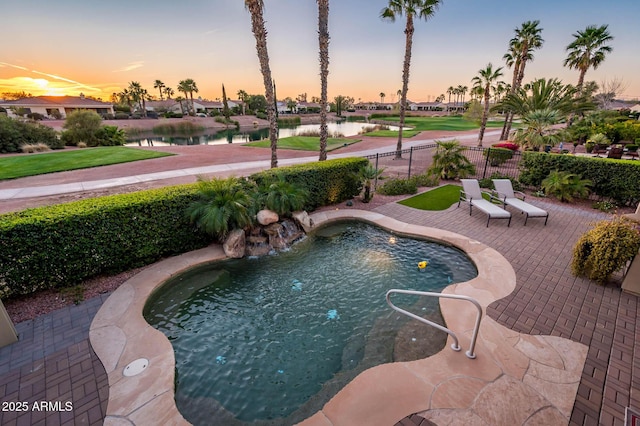 pool at dusk with a patio, a water view, and fence