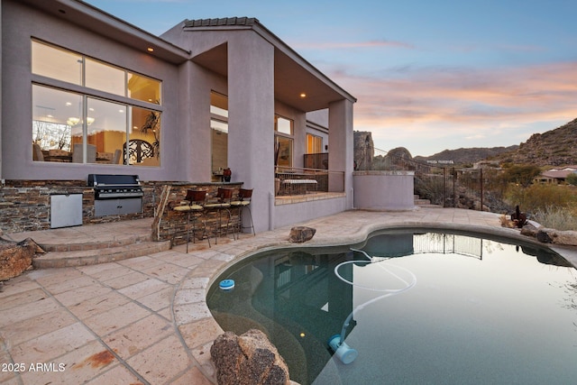 pool at dusk featuring a mountain view, grilling area, an outdoor bar, and a patio area