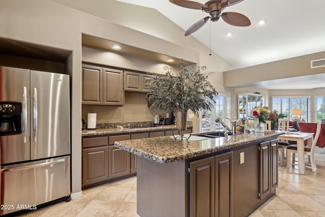 kitchen featuring lofted ceiling, dark stone counters, a kitchen island with sink, a sink, and stainless steel fridge