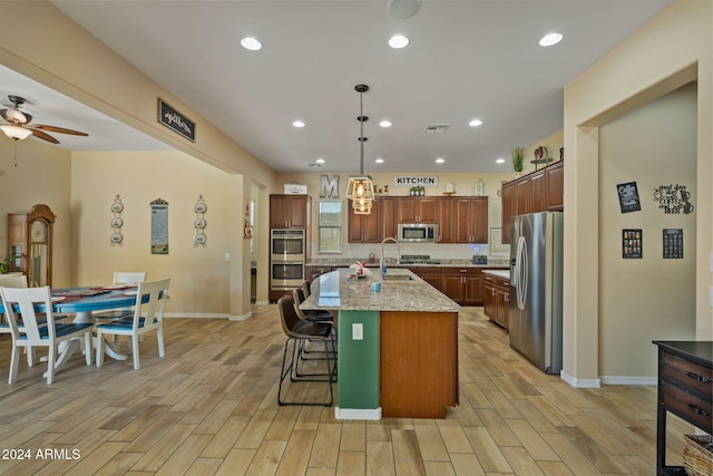 kitchen featuring light stone counters, stainless steel appliances, light hardwood / wood-style flooring, hanging light fixtures, and an island with sink