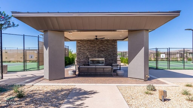 view of patio / terrace with an outdoor stone fireplace, a tennis court, a gate, ceiling fan, and fence