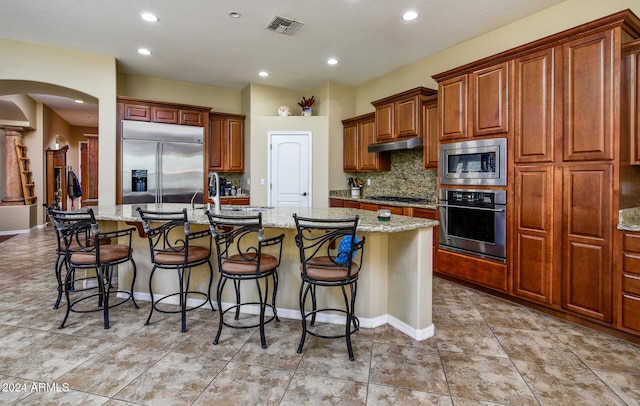 kitchen with light stone counters, built in appliances, tasteful backsplash, an island with sink, and a kitchen breakfast bar