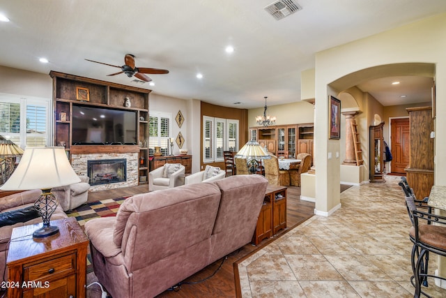 tiled living room with a stone fireplace, a healthy amount of sunlight, and ceiling fan with notable chandelier