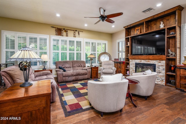 living room featuring a stone fireplace, dark wood-type flooring, a healthy amount of sunlight, and ceiling fan