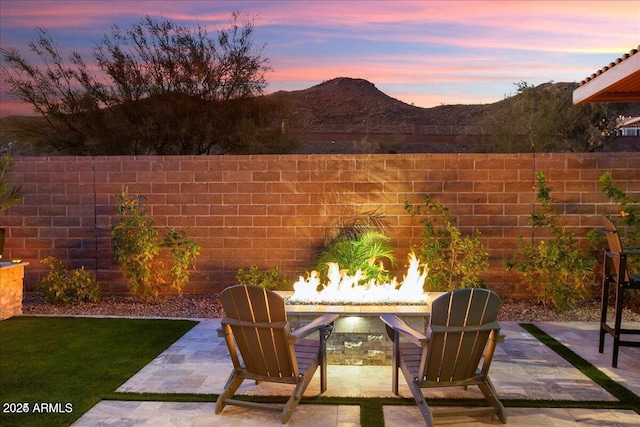 patio terrace at dusk featuring a lit fireplace, a mountain view, and fence