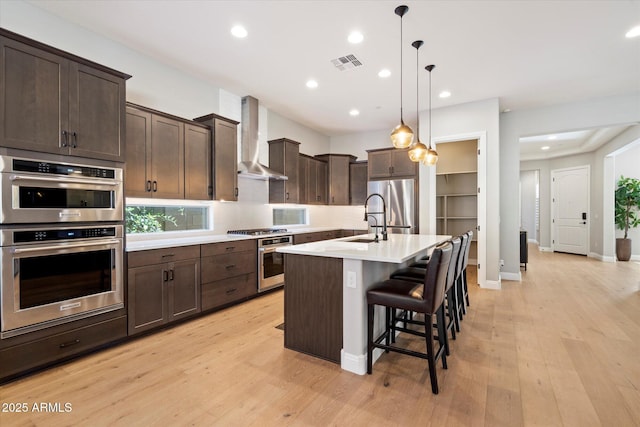kitchen featuring dark brown cabinetry, light countertops, visible vents, wall chimney exhaust hood, and appliances with stainless steel finishes