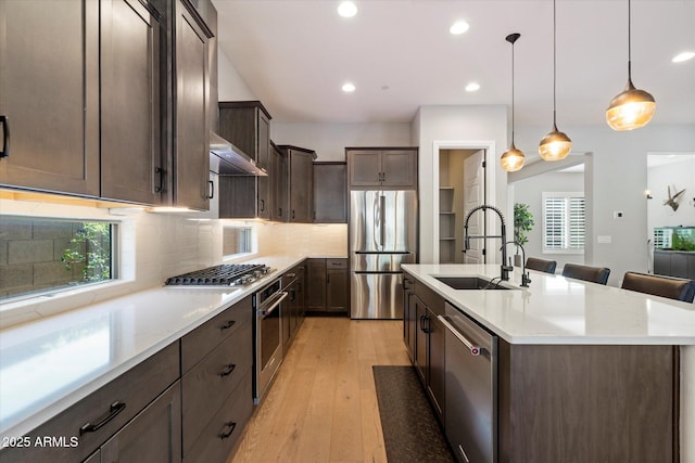 kitchen with light wood-style floors, dark brown cabinets, a kitchen island with sink, a sink, and stainless steel appliances