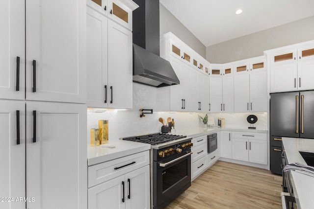 kitchen with high end appliances, wall chimney range hood, light hardwood / wood-style flooring, decorative backsplash, and white cabinetry