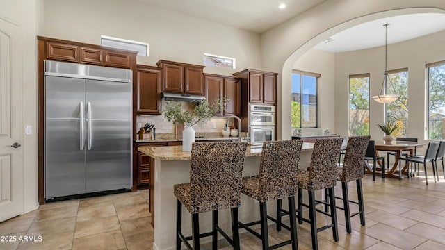kitchen featuring under cabinet range hood, decorative backsplash, arched walkways, an island with sink, and appliances with stainless steel finishes