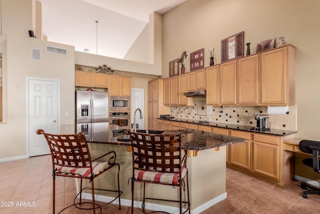 kitchen featuring appliances with stainless steel finishes, a kitchen bar, dark stone countertops, high vaulted ceiling, and a center island with sink