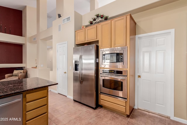 kitchen with light tile patterned floors, appliances with stainless steel finishes, a high ceiling, light brown cabinets, and dark stone counters