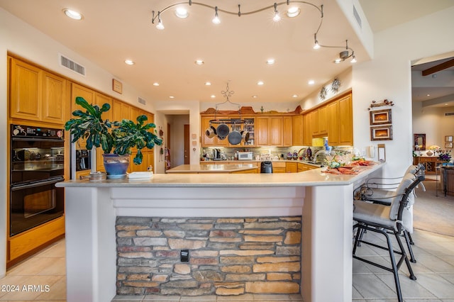 kitchen featuring backsplash, kitchen peninsula, black double oven, a breakfast bar, and light tile patterned flooring