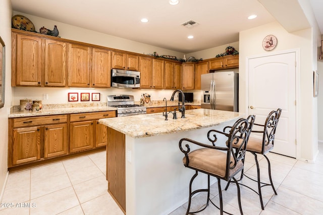 kitchen with visible vents, brown cabinetry, appliances with stainless steel finishes, light stone counters, and a kitchen island with sink