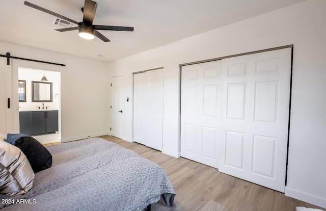 bedroom with ensuite bath, ceiling fan, a barn door, light hardwood / wood-style floors, and multiple closets