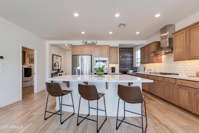 4350 Stage Stop Way, Wickenburg, AZ 85390 - kitchen featuring appliances with stainless steel finishes, wall chimney range hood, washer / clothes dryer, an island with sink, and light wood finished floors