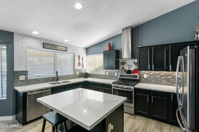 kitchen with sink, light stone counters, appliances with stainless steel finishes, a kitchen island, and wall chimney range hood