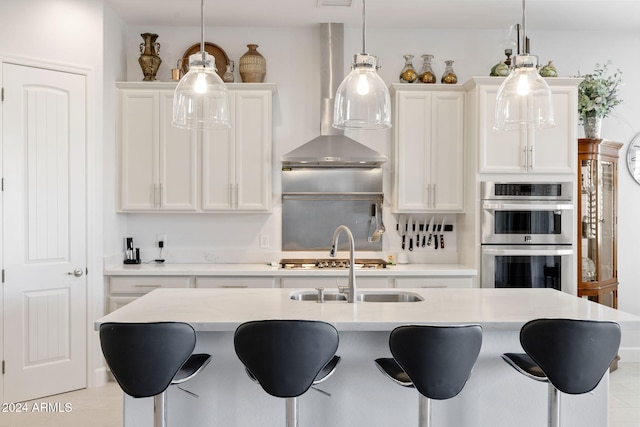 kitchen with decorative light fixtures, a kitchen bar, wall chimney range hood, double oven, and white cabinets
