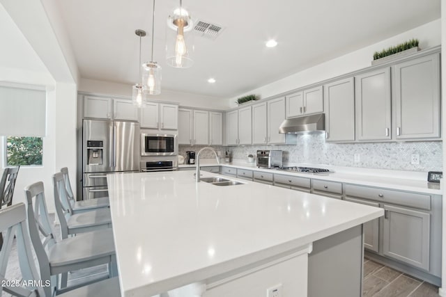 kitchen featuring appliances with stainless steel finishes, a kitchen island with sink, light countertops, under cabinet range hood, and a sink