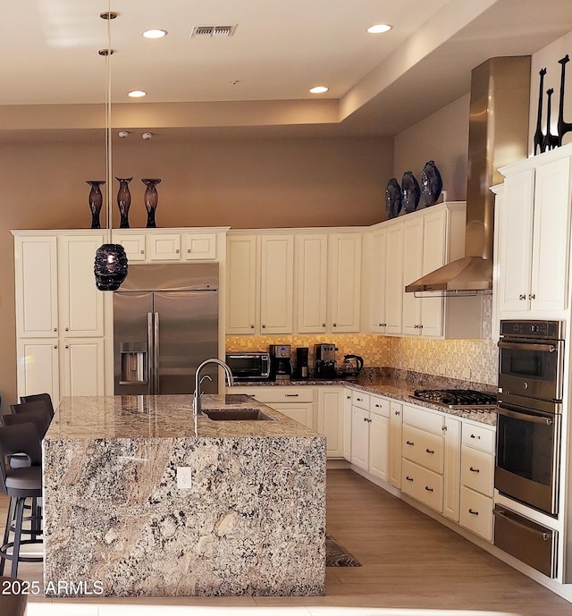 kitchen with wall chimney exhaust hood, a breakfast bar, stainless steel appliances, sink, and an island with sink