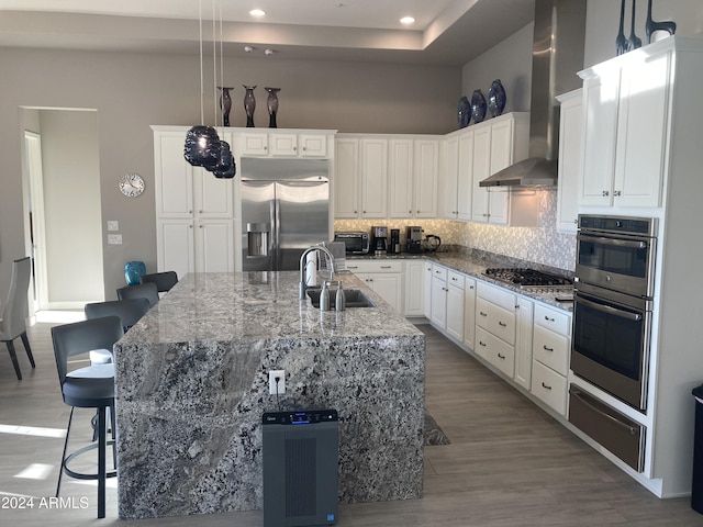 kitchen featuring a center island with sink, hanging light fixtures, wall chimney range hood, and appliances with stainless steel finishes