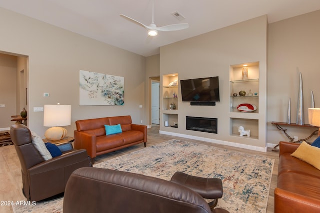 living area with built in shelves, a glass covered fireplace, ceiling fan, and light wood finished floors