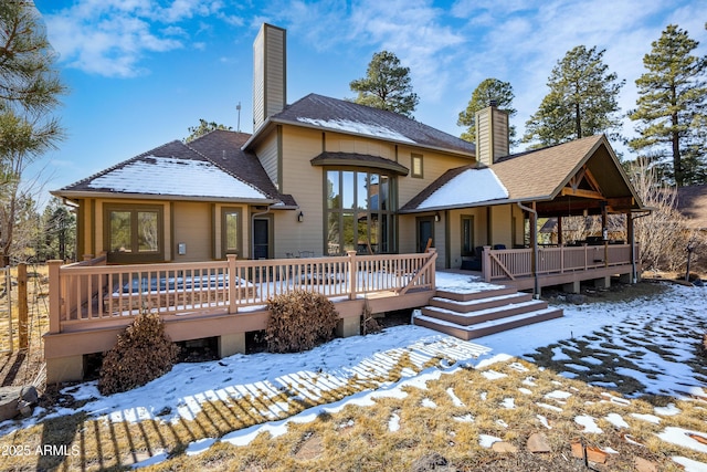 snow covered house featuring a shingled roof, a wooden deck, and a chimney