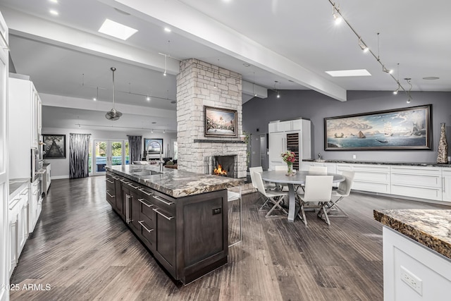 kitchen featuring open floor plan, track lighting, dark brown cabinetry, a stone fireplace, and beamed ceiling