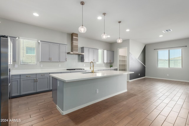 kitchen with decorative light fixtures, stainless steel refrigerator, gas cooktop, a kitchen island with sink, and wall chimney exhaust hood