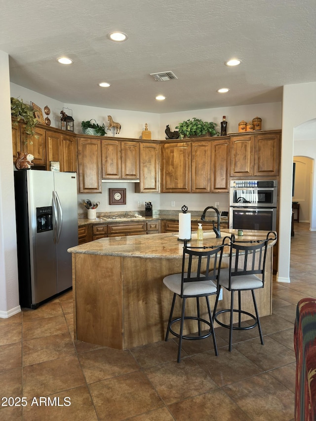 4116 E Sierra Sunset Trl, Cave Creek, AZ 85331 - kitchen with visible vents, brown cabinetry, arched walkways, a kitchen island with sink, and stainless steel appliances