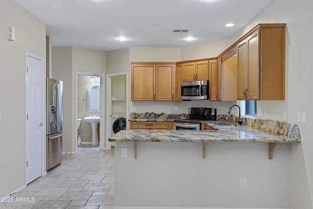 kitchen featuring light stone countertops, appliances with stainless steel finishes, a peninsula, brown cabinetry, and a sink