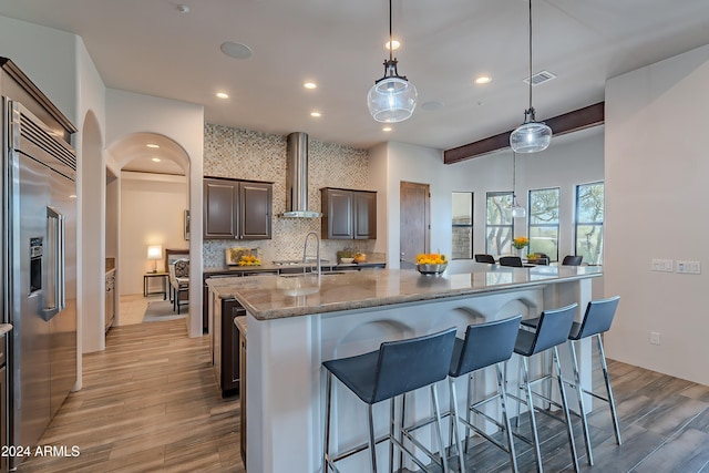 kitchen featuring wall chimney exhaust hood, decorative light fixtures, light hardwood / wood-style flooring, built in fridge, and an island with sink