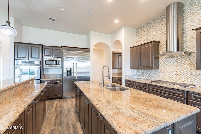 kitchen with wall chimney range hood, sink, built in appliances, an island with sink, and decorative light fixtures