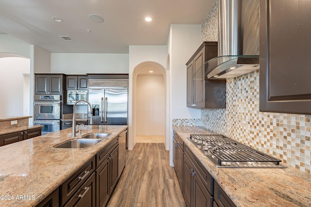 kitchen with wall chimney exhaust hood, light stone countertops, light wood-type flooring, dark brown cabinets, and stainless steel appliances