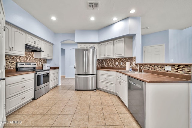 kitchen featuring appliances with stainless steel finishes, under cabinet range hood, a sink, dark countertops, and arched walkways
