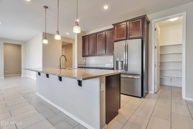 kitchen featuring sink, stainless steel fridge, a kitchen island with sink, hanging light fixtures, and light stone countertops