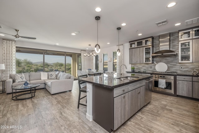 kitchen with hanging light fixtures, sink, a kitchen island with sink, wall chimney exhaust hood, and appliances with stainless steel finishes