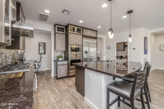 kitchen featuring dark stone countertops, wall chimney exhaust hood, built in appliances, pendant lighting, and light hardwood / wood-style flooring