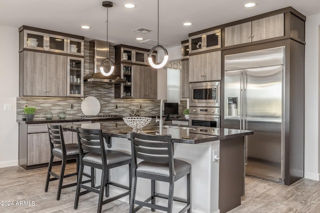 kitchen featuring pendant lighting, a kitchen island with sink, wall chimney exhaust hood, built in appliances, and dark stone countertops