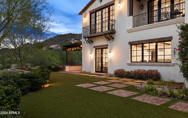 rear view of house with a balcony, stucco siding, a lawn, and french doors