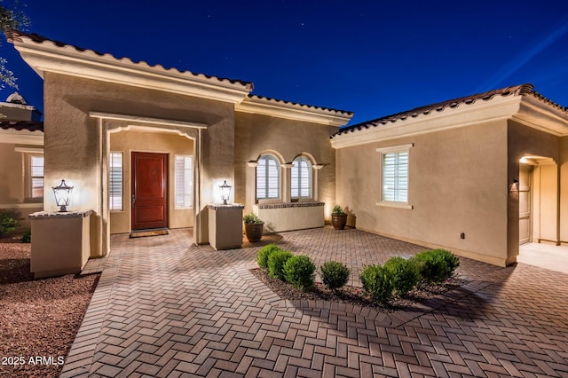 exterior entry at night featuring a patio, a tile roof, and stucco siding