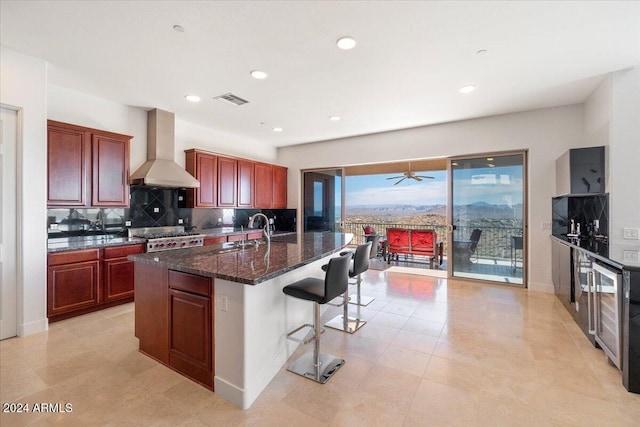 kitchen with dark stone counters, a kitchen island with sink, sink, wall chimney range hood, and a breakfast bar area