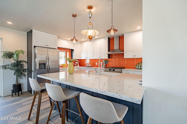 kitchen featuring a sink, wall chimney range hood, appliances with stainless steel finishes, backsplash, and a kitchen bar