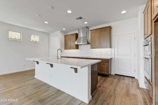 kitchen featuring wall chimney range hood, light wood-type flooring, a breakfast bar area, sink, and an island with sink